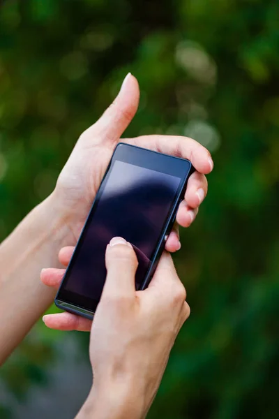 女性の手のサイドビュー夏の緑の背景に親指でダイヤルしながら 現代的なスリックスマートフォンを保持 — ストック写真