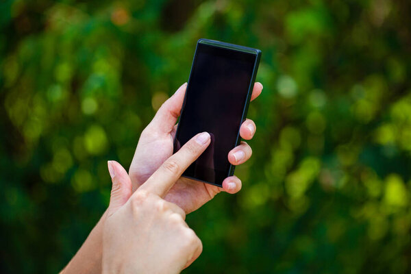 Side view of a woman's hand holding a modern slick smartphone while dialing with her thumb against summer green background