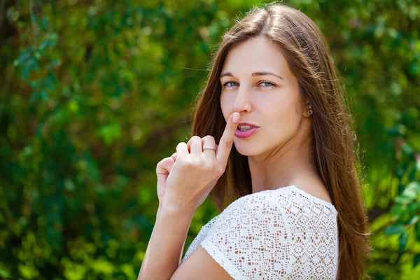 美しいブルネットの若い女性は 緑の夏の庭に対して沈黙の印として唇に人差し指を入れています — ストック写真