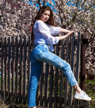 Çiçek açan kiraz ağacının arka planında, kot pantolonlu şık bir genç kızın tüm vücut portresi.