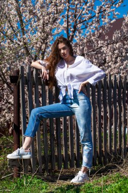 Çiçek açan kiraz ağacının arka planında, kot pantolonlu şık bir genç kızın tüm vücut portresi.