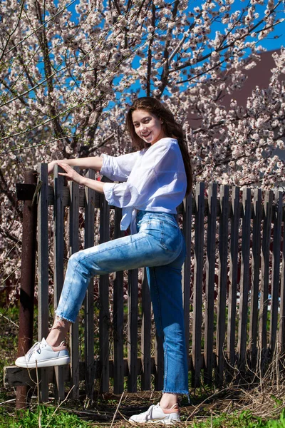 一个穿着蓝色牛仔裤的年轻姑娘的全身像 衬托着盛开的樱桃的背景 — 图库照片