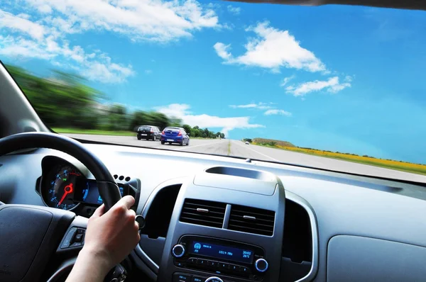 Car dashboard with drivers hand on the black steering wheel against asphalt road and blue sky with clouds