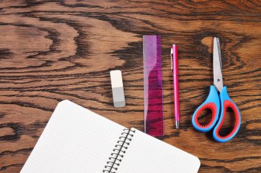 Öğrenci yarıyıl not defteri ile mavi makas, silgi, plastik cetvel ve kahverengi ahşap zemin üzerinde pembe kalem