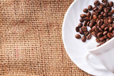 Bir daire bir çuval bezi üzerinde beyaz seramik bardakta üzerinden dökülen kahverengi kahve çekirdekleri