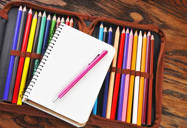 Pencil case full of color pencils with a notebook and pen on the dark brown wooden table