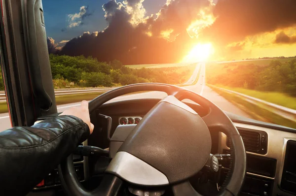 Truck dashboard with drivers hand on the steering wheel on the countryside road against night sky with sunset