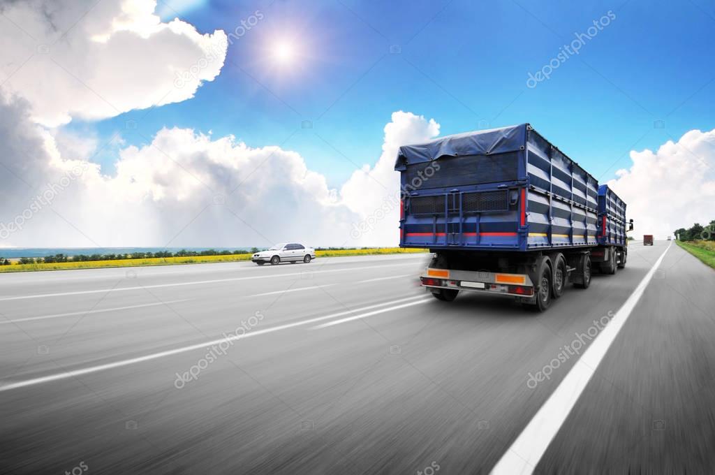 Rear view of big truck driving fast with blue trailer on countryside road against blue sky with clouds