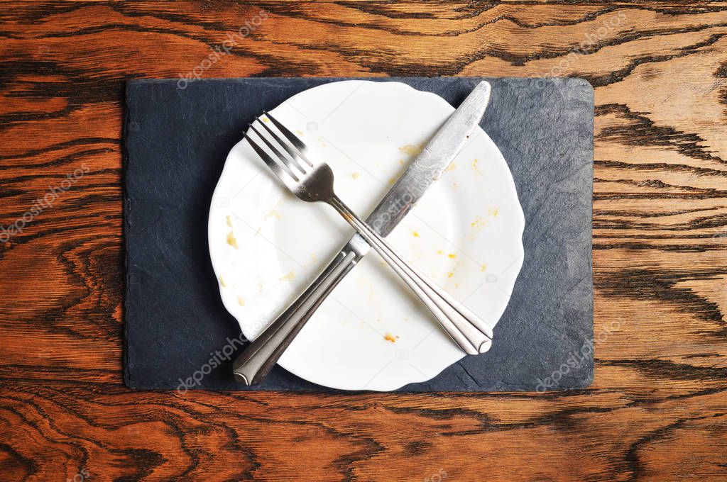 Dirty plate with silver knife and fork crossed on the dark blue board on the wooden background