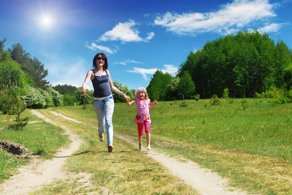 Mother with daughter holding hands and running on the country road with forest on the backround and blue sky with sun and white clouds