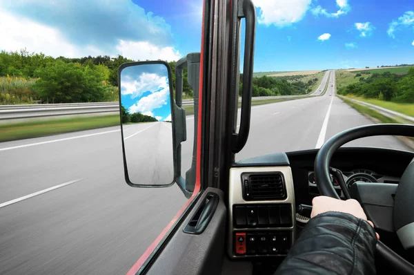Truck dashboard with drivers hand on the steering wheel and side rear-view mirror on the countryside road against blue sky with clouds