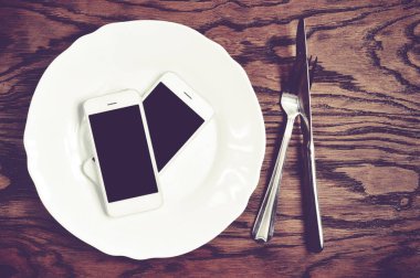 İki plaka üzerinde siyah ekranlı akıllı telefonlar ile gümüş bıçak ve çatal ahşap arka plan üzerinde beyaz