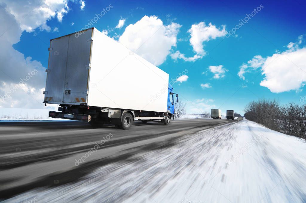 White box truck driving fast on the countryside winter road with snow against blue sky with clouds