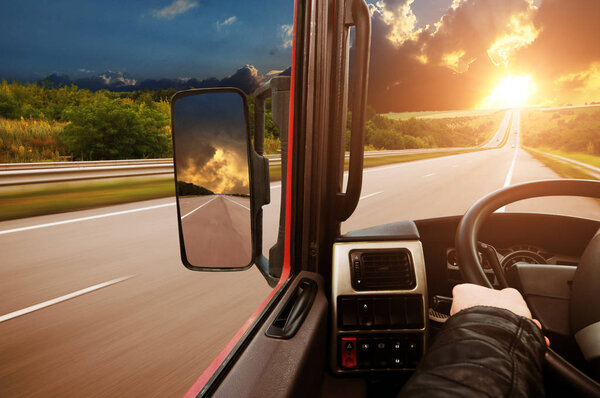 Truck dashboard with drivers hand on the steering wheel and side rear-view mirror on the countryside road against night sky with sunset