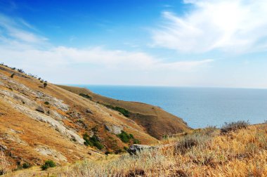 Dağlar ve yeşil çalılar, mavi deniz ve gökyüzü beyaz bulutlar ile kayalık sahil