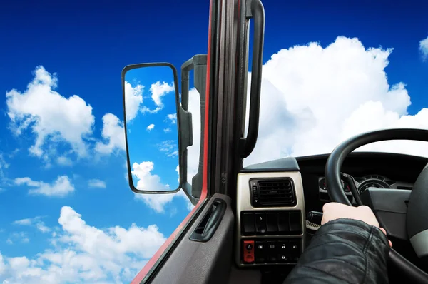Truck dashboard with driver hand on the steering wheel and side rear-view mirror against blue sky with clouds
