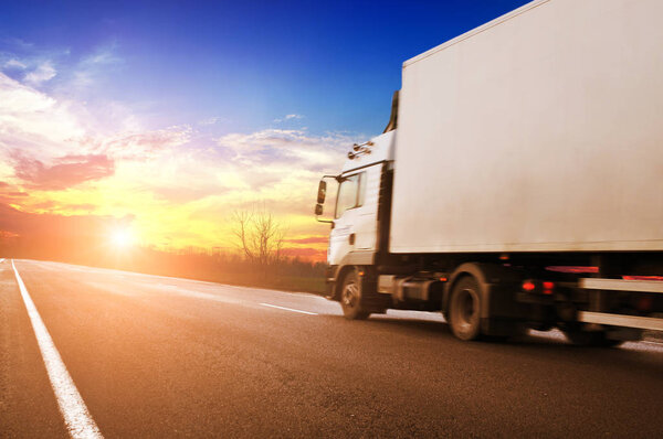 White truck and a white trailer with space for text on the countryside road against blue sky with bright sunset