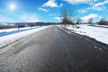 Empty winter countryside road with trees and white snow against blue sky with clouds and sun clipart
