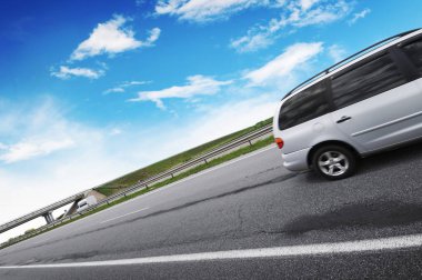 Beyaz bulutlu mavi gökyüzüne karşı asfalt yolda gümüş bir minivan.