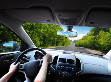Car dashboard with driver hands on the black steering wheel and rear view mirrors on road in motion with green trees against blue sky with clouds