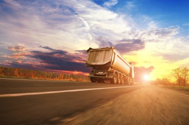 Big white dump truck in motion on the countryside road against sky with sunset