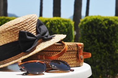 Round straw bag, straw hat and black sunglasses with green bushes and blue sky on the background