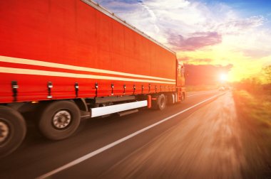 Truck with red trailer with space for text driving fast on the countryside road against sky with sunset