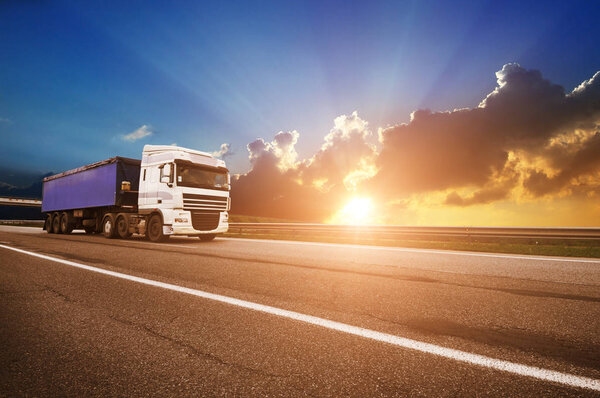 Большой белый грузовик с синим прицепом на сельской дороге против неба с закатом
