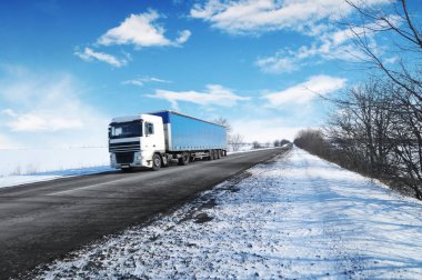 White truck and blue trailer with space for text on the winter countryside road with snow against blue sky with clouds