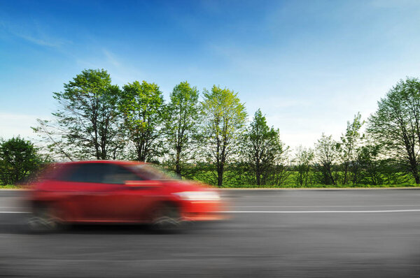 A red hatchback car driving fast on the  countryside road in motion with green trees and bushes against a blue sky
