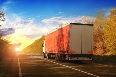 Truck and trailer with space for text on the countryside road with autumn forest against sky with sunset