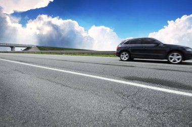 Black crossover car on the countryside asphalt road against blue sky with white clouds