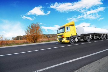Big metal fuel tanker truck shipping fuel on the countryside road with trees against blue sky with clouds