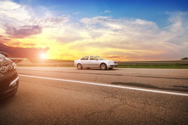 Black and silver sedan cars on the countryside asphalt road with green trees against sky with sunset