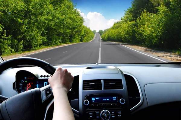 Car dashboard with driver hand on the black steering wheel and road with green trees against blue sky with clouds