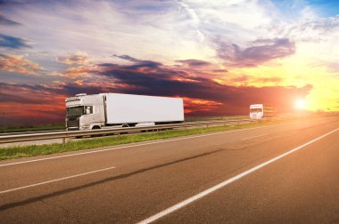 Big truck and white trailer with space for text and other truck on the countryside road against sky with sunset