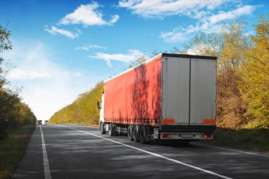 Truck and trailer with space for text on the countryside road with autumn forest against blue sky with clouds