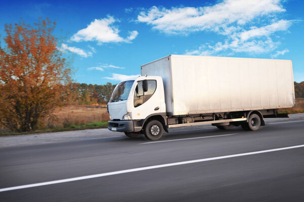 White boxed truck driving fast on the countryside road with trees and bushes against blue sky with clouds