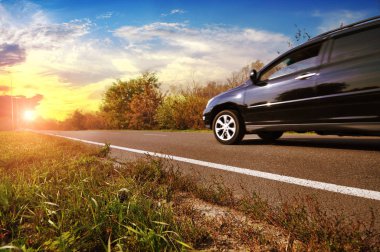 Siyah aile arabası asfalt yolda, gökyüzüne karşı yeşil ağaçlar ve gün batımı.