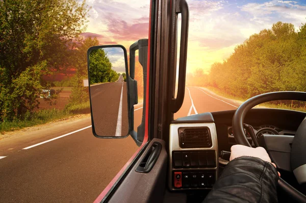 Truck dashboard with driver hand on the steering wheel and side rear-view mirror on the countryside road against sky with sunset