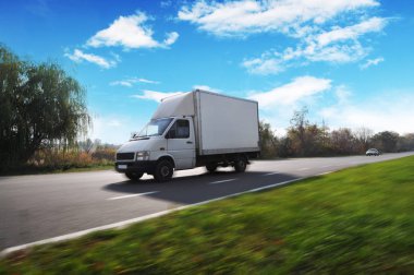 Kırsal yolda hızlı giden beyaz kamyonet yeşil ağaçlar ve çalılarla mavi gökyüzüne doğru bulutlarla ilerliyor.