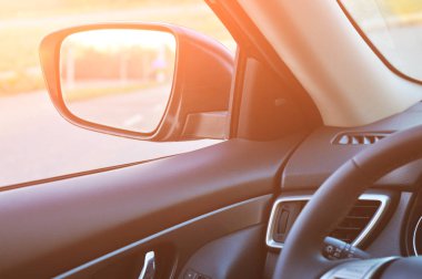 Siyah bir arabanın içi, dikiz aynası ve güneş ışığıyla bulanık kırsal yol.