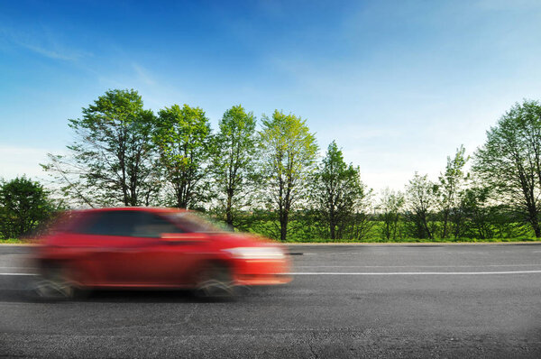Red hatchback car driving fast on the countryside road with green trees and bushes against blue sky
