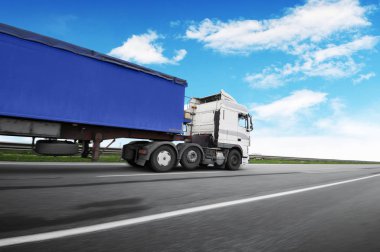 Big white truck and blue trailer with space for text driving fast on the countryside road against blue sky with clouds