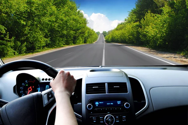 Car dashboard with driver hand on the black steering wheel and road in motion with green trees against blue sky with clouds