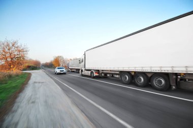 Büyük kamyonet ve beyaz römork. Mavi gökyüzüne karşı kırsal yolda hızla ilerleyen araçlar.