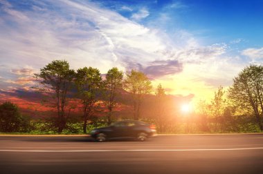 Black sedan car driving fast on the countryside asphalt road in motion with green trees against sky with sunset clipart
