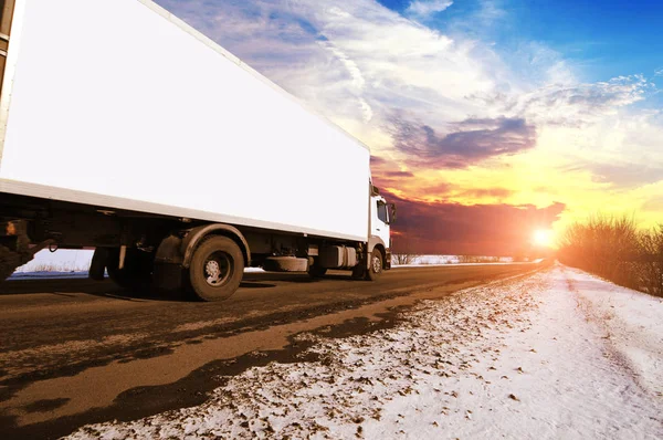 White box truck with space for text on the countryside winter road with snow against sky with sunset