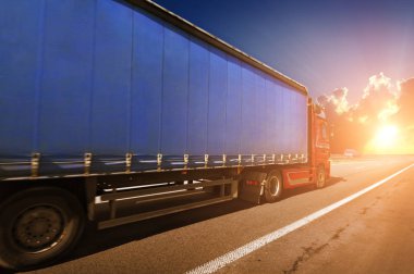 Büyük kırmızı kamyon ve mavi römork kırsal yolda gün batımında gökyüzüne karşı yazı için yer var.