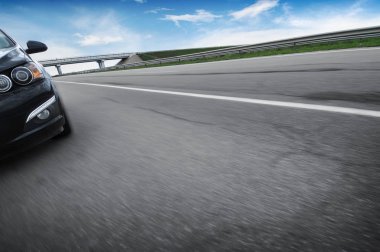 Siyah aile arabası asfalt yolda hızla ilerlerken mavi gökyüzüne karşı bulutlarla köprü kuruyor.